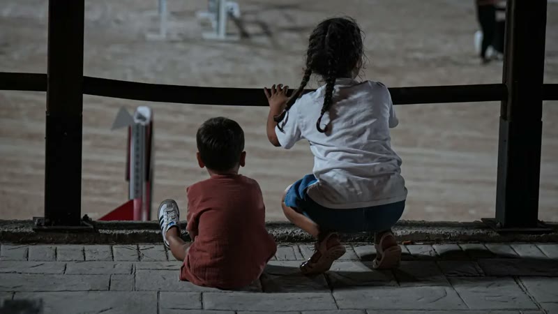 Manej kenarında yere oturmuş, yarışmayı izleyen iki çocuk, arkadan