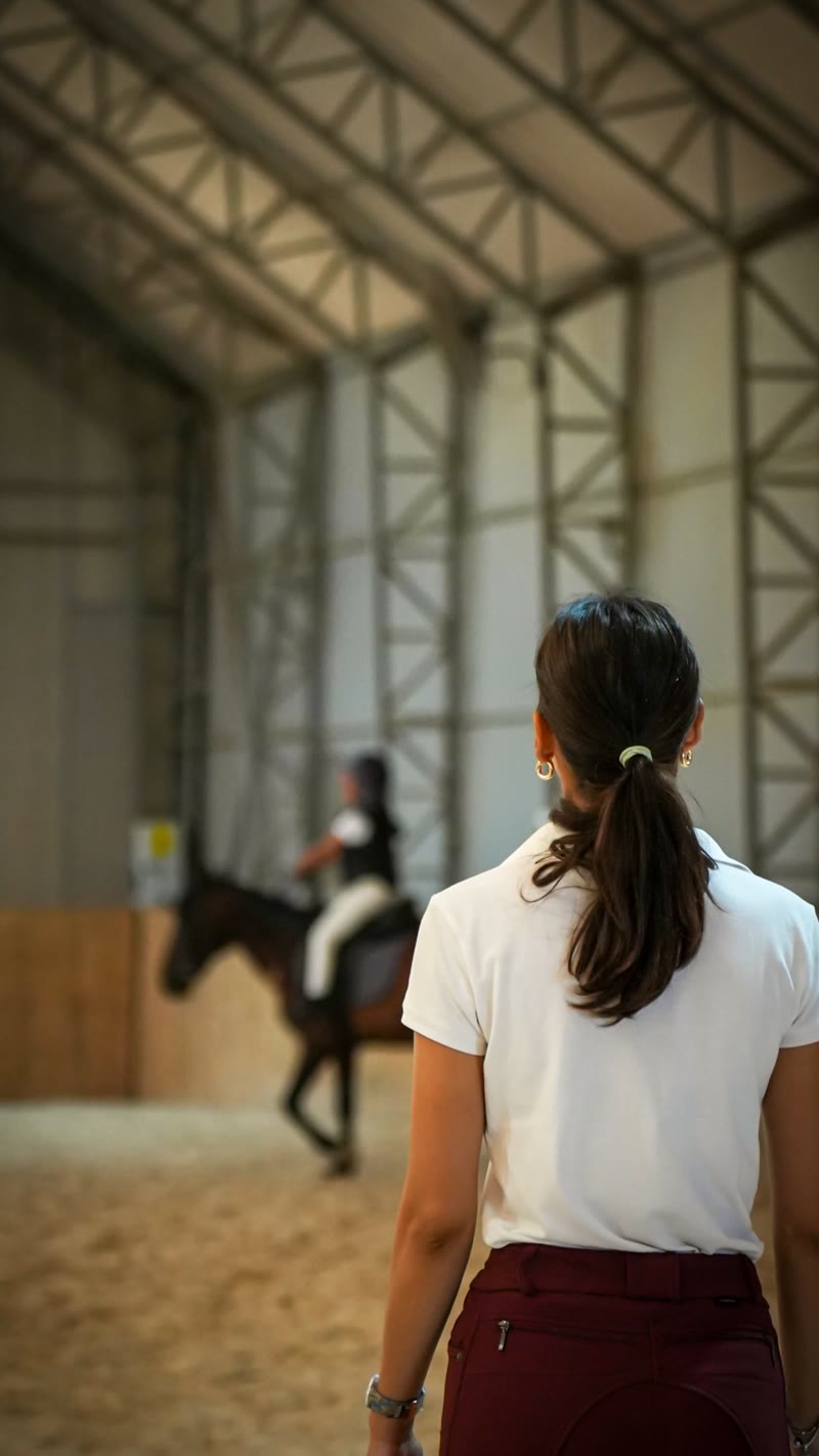 Kapalı manejde arkası dönük duran eğitmen, ileride pony üstünde bir çocuk